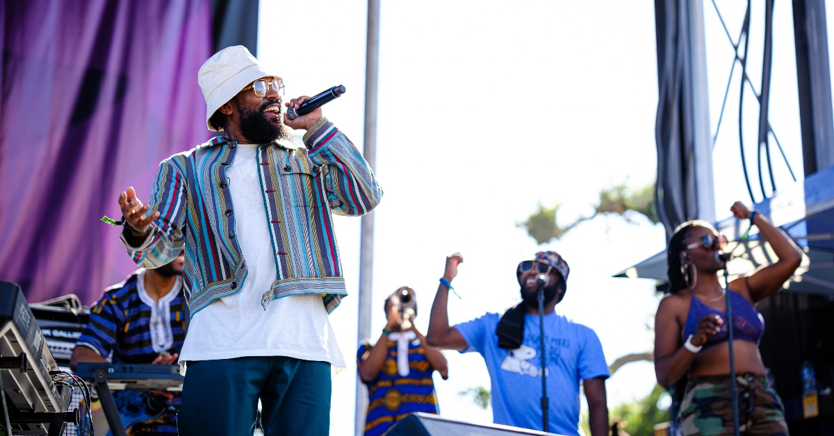 PJ Morton wears a white bucket hat and a multi-colored jacket and sings on stage with a band.